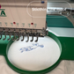 Embroidery machine stitching a design on fabric with 'teememento' branding.